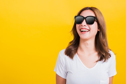 Portrait Asian Thai beautiful young woman happy smiling white teeth with sunglasses and looking side space, studio shot isolated on yellow background, with copy space