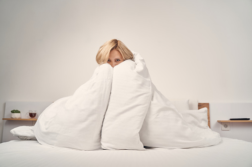 Front view of a young Caucasian female wrapped in a duvet sitting on her bed