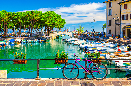 Bicycle bike near fence of old harbour Porto Vecchio with motor boats on turquoise water and Venetian bridge in historical centre of Desenzano del Garda town, blue sky, Lombardy, Northern Italy
