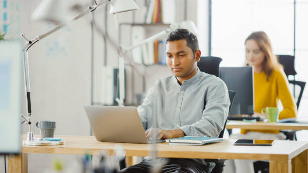 istock Smart and Handsome Indian Information Technology Specialist Sitting at His Desk works on a Laptop. In the Background Modern Office with Diverse Team of Young Professionals Working. 1248743495