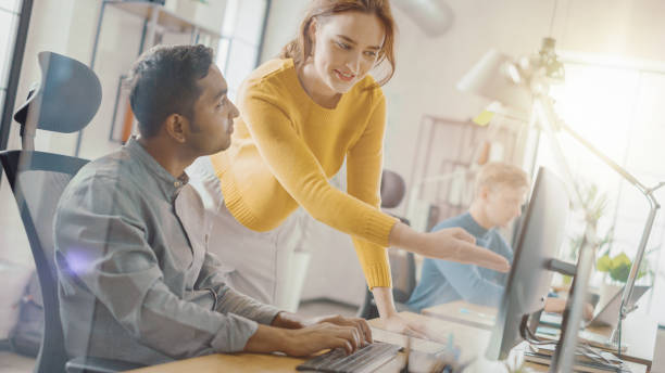 istock Handsome Indian Professional Sitting at His Desk Working on Laptop Computer, Young Beautiful Team Leader Gives Advice about Project Details. Office with Young Professionals Work. Dutch Angle. 1248743506