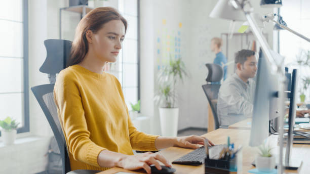 istock Beautiful and Creative Young Woman Sitting at Her Desk Using Laptop Computer. In the Background Bright Office where Diverse Team of Young Professionals Work. 1248743531