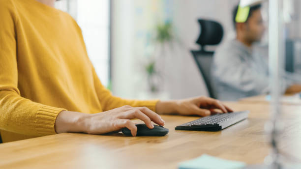 giovane donna anonima seduta alla sua scrivania usando il computer portatile. concentrati sulle mani usando mouse e tastiera. nell'ufficio luminoso di sfondo dove lavorano diversi team di giovani professionisti. - single click foto e immagini stock