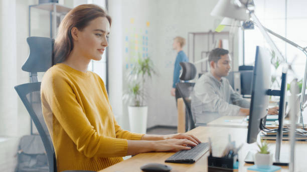 istock Beautiful and Creative Young Woman Sitting at Her Desk Using Laptop Computer. In the Background Bright Office where Diverse Team of Young Professionals Work. 1248743558