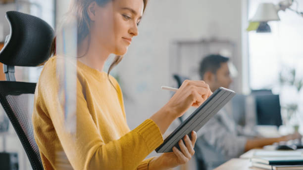 Talented Female Designer Sitting at Her Desk She's Drawing, Writing and Using Pen with Digital Tablet Computer. Focus on Hands with Pen. Bright Office where Diverse Team of Young Professionals Work.