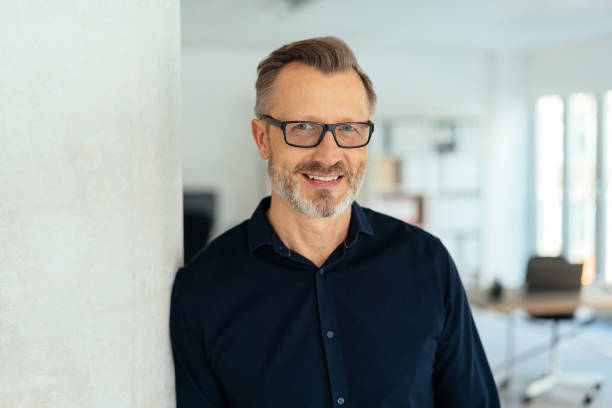 Smiling handsome middle-aged man in black shirt Indoor close-up portrait of a smiling handsome bearded middle-aged man in glasses and black shirt, leaning with his shoulder on white wall and looking at camera with friendly face, standing in office mid adult stock pictures, royalty-free photos & images