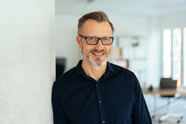 Smiling handsome middle-aged man in black shirt Indoor close-up portrait of a smiling handsome bearded middle-aged man in glasses and black shirt, leaning with his shoulder on white wall and looking at camera with friendly face, standing in office mature businessman stock pictures, royalty-free photos & images