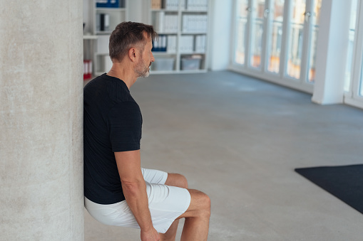 Fit healthy man doing a wall leg sit or squat in an office gym to strengthen and tone his muscles in an active lifestyle concept with copy space