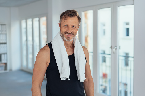 Fit athletic man after a workout in a gym standing with a towel draped around his neck smiling at the camera