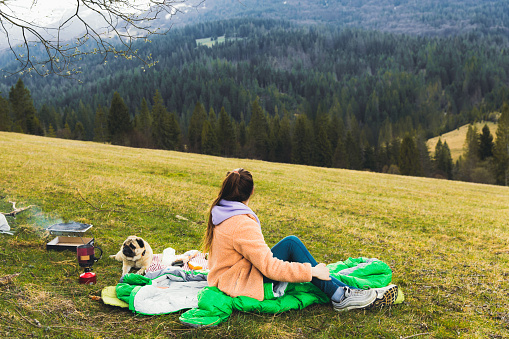 Woman traveler with long hair and a small cute dog - pug breed camping on the beautiful fresh spring meadow in a sleeping bag, having picnic near the lonely tree with view of the snowcapped Carpathian Mountains, Ukraine