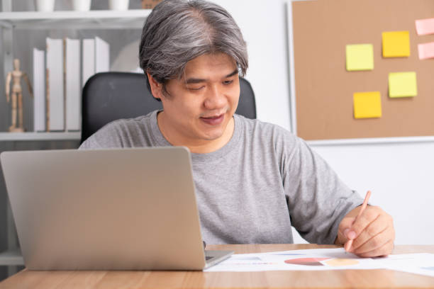 istock Smiling Asian businessman working on the computer and take note on notebook. Concept of working from home until Coronavirus (COVID-19) quarantine, happily freelance working lifestyle 1248760353
