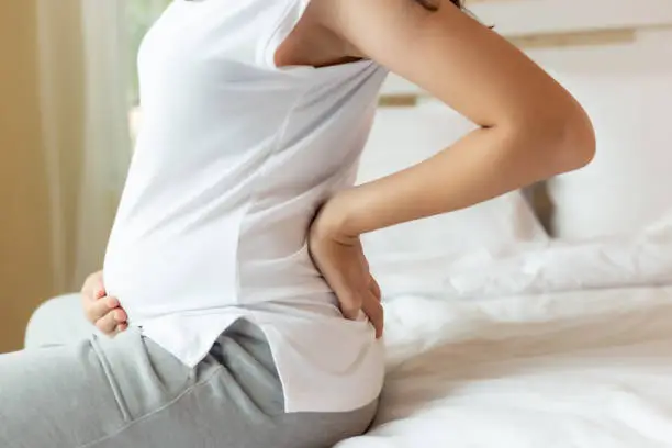 Photo of Pregnant woman touching tummy and suffering from back ache sitting on bed at home. Young attractive pregnant mother get back pain, her fetus or baby and belly bigger than before that mom get painful