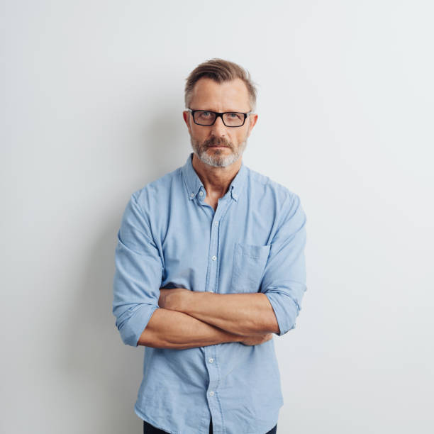 Stern serious businessman staring at the camera Stern serious businessman staring intently at the camera with a deadpan expression and folded arms against a white studio background in square format uncompromising stock pictures, royalty-free photos & images