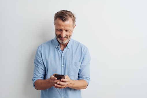 Smiling middle-aged man texting a message on his handheld mobile phone over a white studio background with copy space