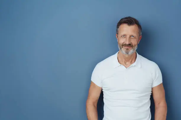 Casual bearded middle-aged man in a casual white t-shirt standing smiling at the camera against a blue studio background with copy space