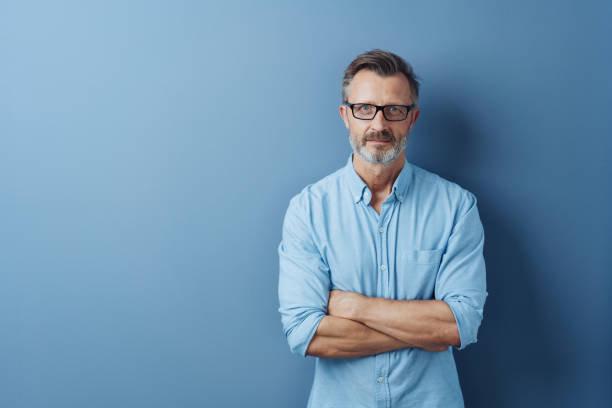 Serious authoritative man with folded arms Serious authoritative bearded middle-aged man with folded arms standing staring intently at the camera against a blue studio background with copy space grave stock pictures, royalty-free photos & images