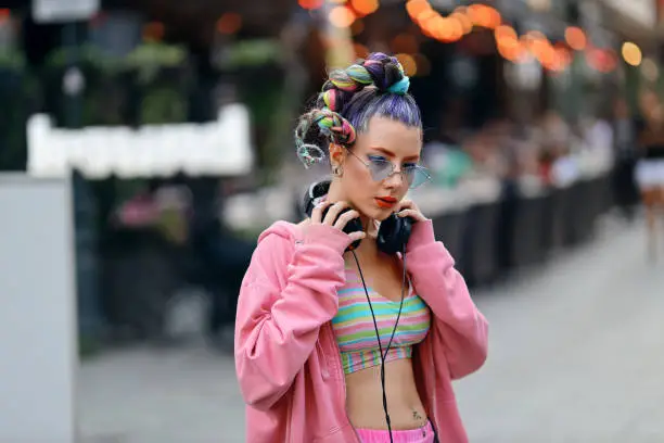 Fashion cool funky girl in headphones listening to music wearing colorful pink sweater and fancy sunglasses on the streets with pubs and bars in city downtown