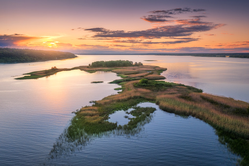 Aerial view of beautiful small islands with green trees and grass on the river at sunset in summer. Colorful landscape with island, meadows, orange sky reflection in blue water. Top view. Nature
