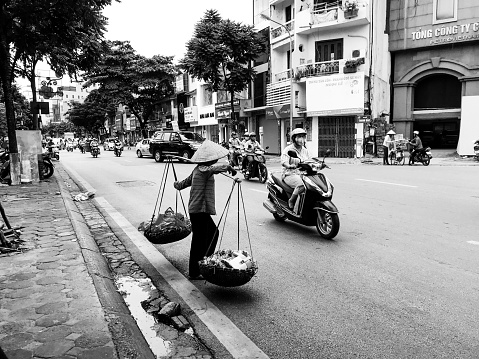 Traditional Vietnamese woman prepares to cross snarling Hanoi traffic while balancing goods, shot in black and white
