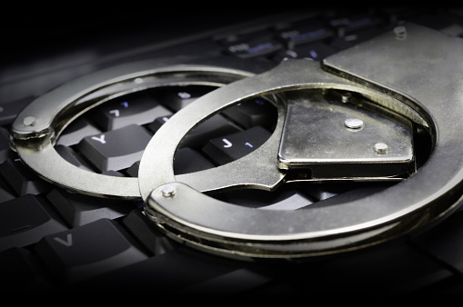 A conceptual image with steel law enforcement handcuffs placed on top of a dark keypad for online legal systems in order.