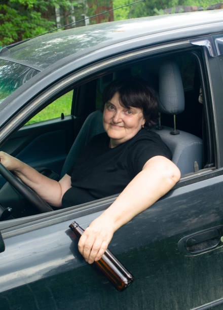 飲酒運転。車の車輪に座って彼女の手にボトルを持つ酔っ払った高齢の女性 - eastern european caucasian one person alcoholism ストックフォトと画像