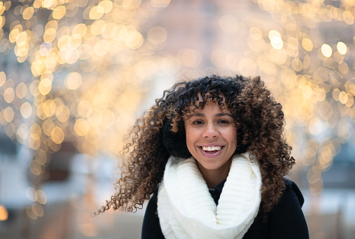 A woman of colour is outside in the winter as shown by Christmas lights. She is smiling at the camera.