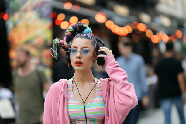 Fashion cool funky girl in headphones listening to music wearing colorful pink sweater and fancy sunglasses on a street with pubs and bars. Unrecognisable incidental people behind stock photo