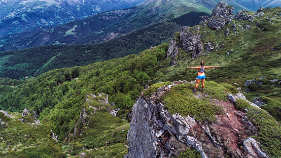 Young woman relaxing and enjoying the view during hard trail running training on the top of mountain. She wears sport clothing and standing on top of mountain with arms outstretched.