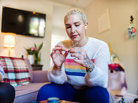 Young blonde woman with tattoos rolling up a joint of recreational marijuana. Interior of small apartment indoors.