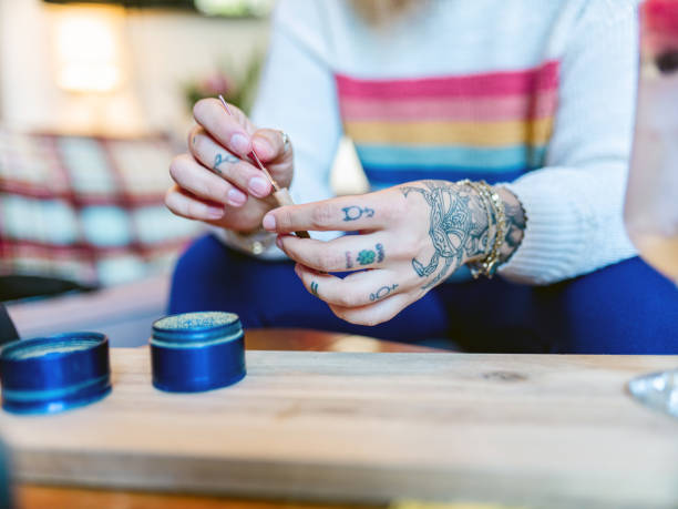 Hands of Young woman rolling up a joint of marijuana Close up of hands of Young woman with tattoos rolling up a joint of recreational marijuana. Interior of small apartment indoors. marijuana tattoo stock pictures, royalty-free photos & images