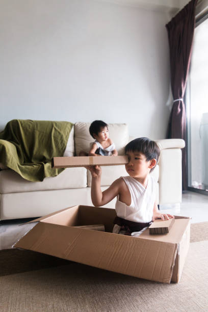 istock Boy sails cardboard box ship; brother sits on couch 1248803245