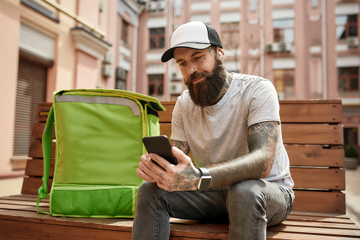 Brutal bearded delivery man in cap with thermo bag or backpack checking order using smartphone, sitting on the bench outdoors. Courier, delivery service concept. Horizontal shot