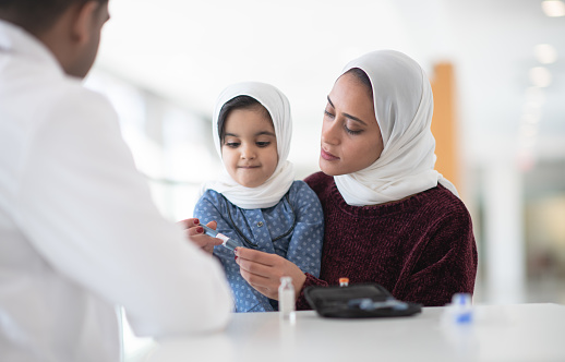 A doctor of color talks with woman who is wearing a hijab, and her daughter or 3 or 4 years old who is sitting in her lap.