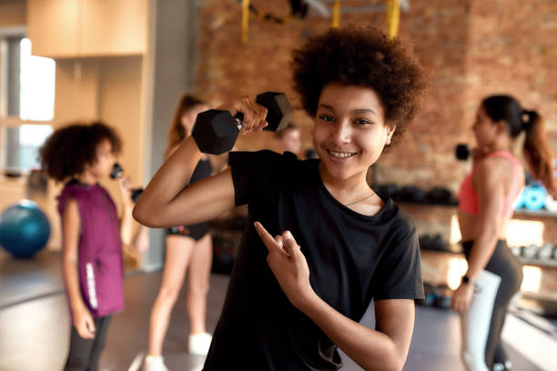 istock African american boy showing muscle while exercising using dumbbell in gym together with female trainer and other kids. Sport, healthy lifestyle, physical education concept 1248806706