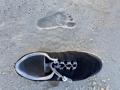 High angle closeup photo of a child’s small footprint in the sand on and beach, and next to it an adult’s empty black and white sports shoe.