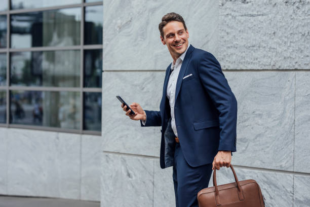 Smiling Handsome Businessman in a Blue Suit Checking Social Media on his Smartphone on the Street Technology on the go: a happy businessman using his mobile phone on the street. man looking at phone over shoulder stock pictures, royalty-free photos & images