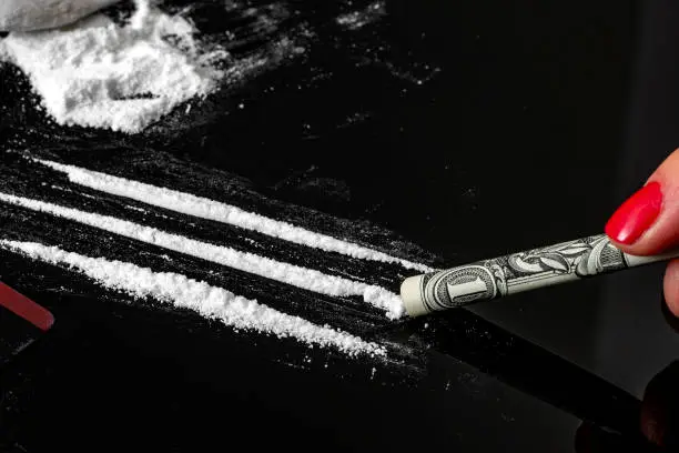 Drug addiction and substance abuse concept theme with close up on a troubled addict using a one dollar bill as a straw to snort a line of cocaine on a dark mirror table next to a pile of white powder