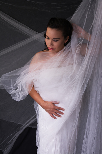 Vertical studio shot on black with sheer white draping of pregnant Native woman touching belly and looking down and to side