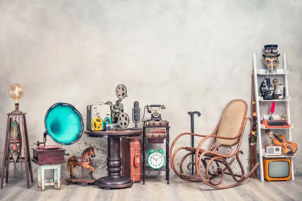 Photo of Antique gramophone, rocking chair, old typewriter, retro radio, tape recorder, projector, books, clock, camera, fiddle, mask, cylinder hat, cane, suitcase, bow. Vintage style filtered photo