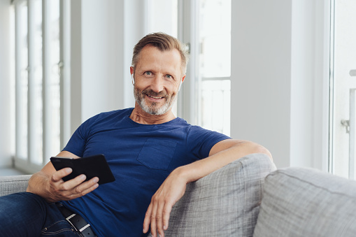 Relaxed attractive middle-aged man on a sofa sitting listening to music on his mobile phone using an earbud as he smiles at the camera