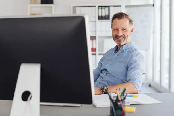 Smiling friendly relaxed businessman in the office seated at his desk behind a desktop monitor looking attentively at the camera