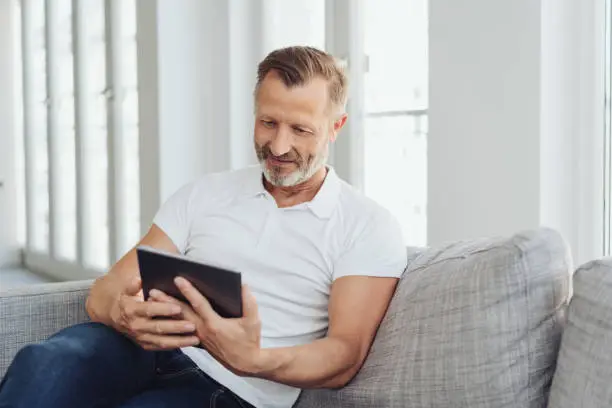 Man relaxing at home on a comfortable sofa with his tablet pc reading online with a quiet smile of pleasure