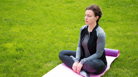Yoga fitness caucasian woman wearing sportswear practicing yoga,closed eyes outdoor in park on green grass.Calm athlete meditating before strength training workout sitting in lotus position on mat