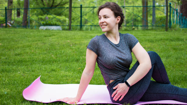 Yoga stretch exercise fit happy caucasian young woman stretching lower back for spine health in city park on green grass.Athletic girl sitting on fitness mat training class outdoor.Seated spinal twist ejercicio elástico de yoga ajuste mujer joven caucásica feliz que se extiende en la parte inferior de la espalda para la salud de la columna vertebral en el parque de la ciudad en hierba verde. chica atlética sentada en la clase de entrenamiento de este