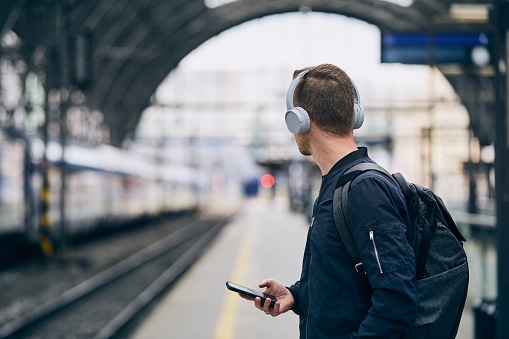 Young man with headphones listening music and waiting for train at railroad station.