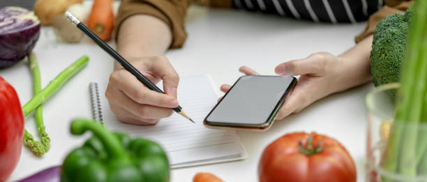istock Female noting ingredient on notebook while looking information on mock-up smartphone on kitchen table with fresh vegetables 1248891610