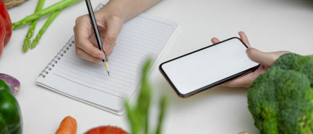 istock Female noting diet plan while looking information on mock-up smartphone on kitchen table with fresh vegetables 1248891657