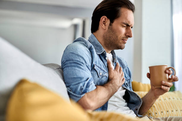 Young man having a chest pain while drinking tea at home. Young man feeling sick and holding his chest in pain while drinking tea in the living room. chest pain stock pictures, royalty-free photos & images