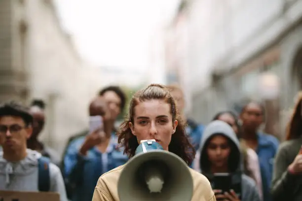 Young woman with a megaphone with group of demonstrator in background. Woman protesting with megaphone in the city.