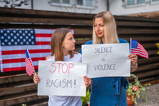 Young caucasian woman with a handwritten message and a little girl holding small american flags and white posters with handwritten message: STOP RACISM! and SIILENCE IS VIOLENCE. They are standing outdoors and they are looking at camera. American flag on the wooden fence in the blurred background.